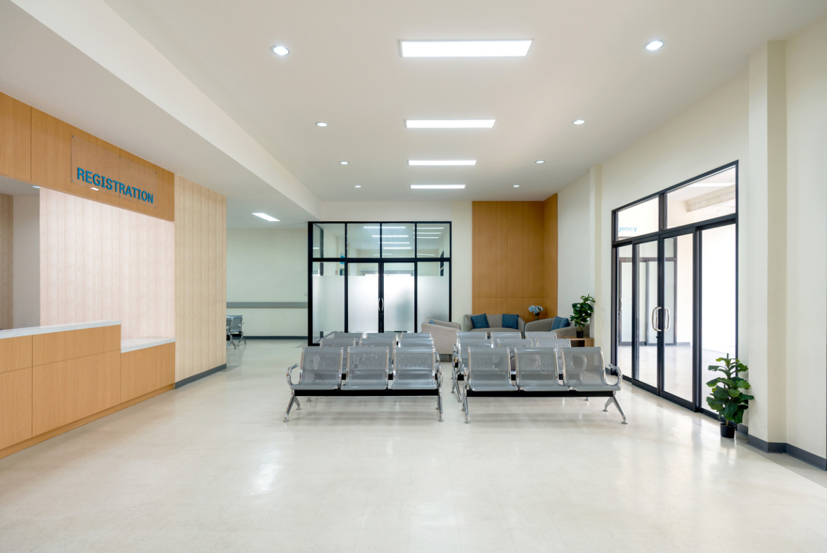 Waiting area in a hospital. Patient or visitor can check in for appointment or service. There are rows of connected grey seating for individual waiting. Glass door on the right lead to emergency area.